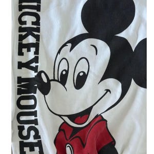 May include: White t-shirt featuring a large graphic of Mickey Mouse in black, red, and white. The text "MICKEY MOUSE" is printed vertically on the left side of the shirt. The cartoon character is smiling.
