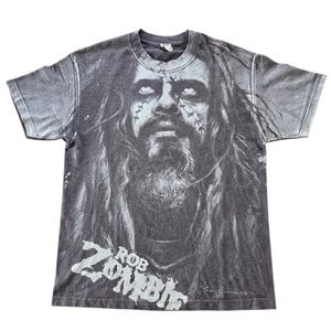 May include: A gray t-shirt featuring a black and white portrait of Rob Zombie. The image is centered on the shirt and includes the text "Rob Zombie" in white block letters. The shirt has a vintage, faded appearance.
