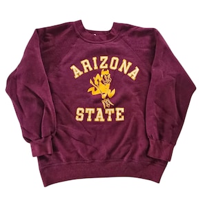 以下が含まれることがあります： 金色の文字とマスコットグラフィックが入った、マルーン色のArizona Stateのスウェットシャツ。「ARIZONA STATE」の文字がブロックレタースタイルで入っています。マスコットは悪魔のような姿です。