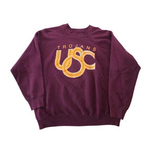 May include: A maroon long-sleeved crewneck sweatshirt featuring the text TROJANS and the letters USC in a stylised yellow and white graphic print on the chest. The garment has raglan sleeves, a ribbed collar, ribbed cuffs, and a ribbed hemline.