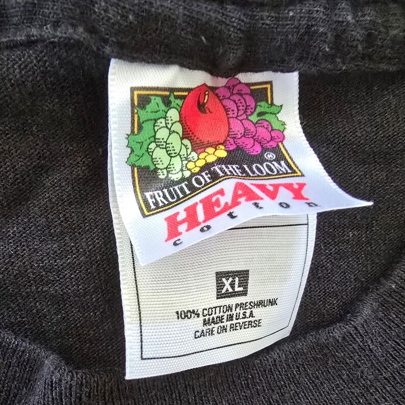May include: Close-up of a black t-shirt with a Fruit of the Loom tag. The tag features a logo with a red apple, purple grapes, and green leaves. The tag also includes the text "HEAVY COTTON" and "XL".