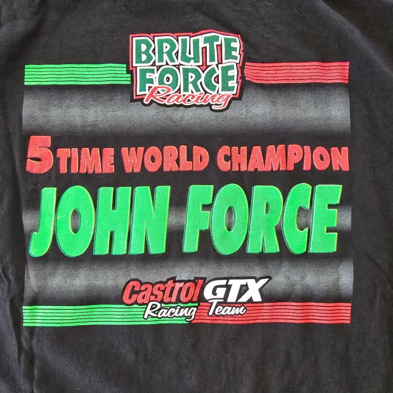 May include: Black t-shirt with racing graphics. The shirt features the text "BRUTE FORCE Racing" and "5 TIME WORLD CHAMPION JOHN FORCE". Below is "Castrol GTX Racing Team". Green and red accents are used throughout the design.