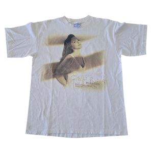 May include: White t-shirt featuring a sepia-toned image of Gloria Estefan. The graphic includes the name "Gloria Estefan" and the text "evolution tour '96". The shirt has short sleeves and a crew neck.