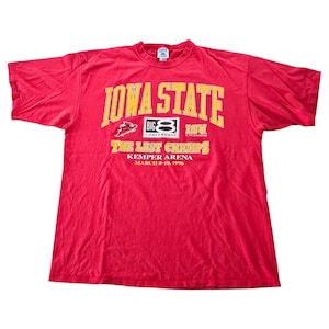 Può includere: Una maglietta rossa degli Iowa State Cyclones con scritte gialle. La maglietta presenta la scritta "IOWA STATE" sopra "BIG 8 CONFERENCE" e "THE LAST CHAMPS" con "KEMPER ARENA MARCH 8-10, 1996" sotto.