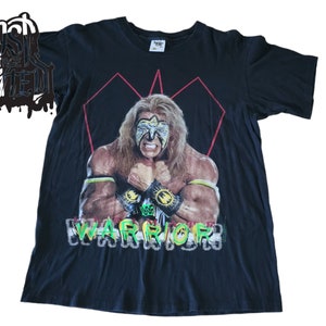 May include: Black t-shirt with a graphic print of a wrestler with a yellow and black face paint. The wrestler is wearing a yellow and black wrestling outfit. The text "Warrior" is printed in green and yellow letters below the wrestler's image.