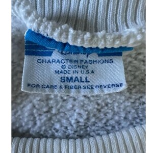 May include: Close-up of a white sweatshirt tag with blue accents. The tag reads "Disney Character Fashions, Made in U.S.A., SMALL." The sweatshirt has ribbed cuffs and a textured fabric.