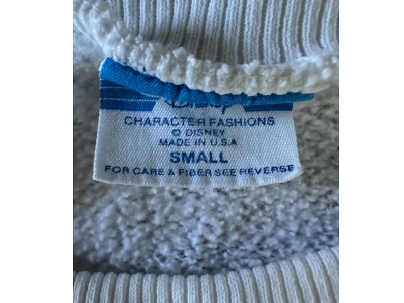 May include: Close-up of a white sweatshirt tag with blue accents. The tag reads "Disney Character Fashions, Made in U.S.A., SMALL." The sweatshirt has ribbed cuffs and a textured fabric.