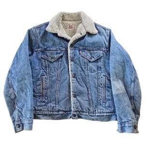 May include: A light blue denim jacket with a cream-colored, fuzzy shearling collar. The jacket has two chest pockets, two side pockets, and a button-up front. The jacket has a classic, vintage style.