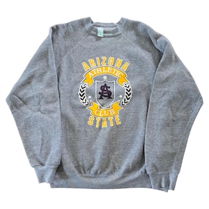 Op de afbeelding: Een grijze crewneck sweatshirt met een afbeelding op de voorkant. De afbeelding toont de woorden "ARIZONA ATHLETIC CLUB STATE" in geel en wit, met een zwart en goud embleem in het midden. De sweatshirt heeft een vintage look.