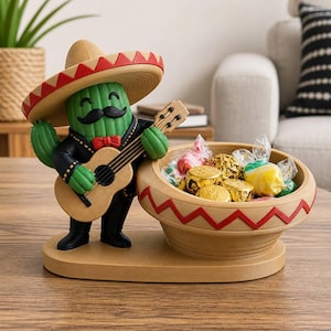 May include: A green cactus figurine wearing a tan sombrero with a red zigzag pattern, a black jacket, and a red bowtie. The cactus holds a tan guitar and stands next to a matching tan bowl with a red zigzag pattern filled with wrapped sweets.