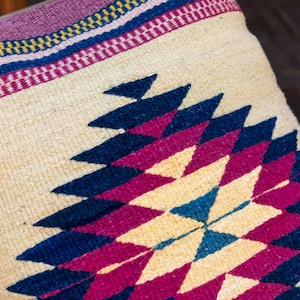 May include: A decorative pillow with a geometric pattern in shades of pink, blue, and cream. The pillow is woven with a traditional design and has a fringe border.