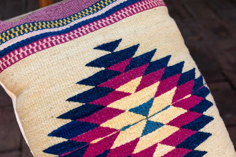 May include: A decorative pillow with a geometric pattern in shades of pink, blue, and cream. The pillow is woven with a traditional design and has a fringe border.