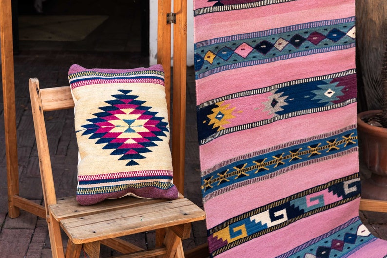 May include: A wooden chair with a colorful patterned pillow on the seat. The chair is in front of a pink and blue patterned rug.