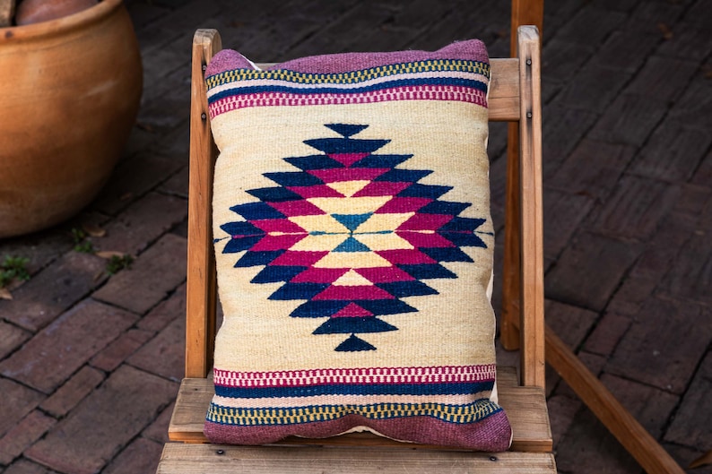 May include: A beige throw pillow with a geometric pattern in shades of pink, blue, and black. The pillow is sitting on a wooden chair.