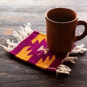 May include: A brown clay mug sits on a colorful woven coaster with fringe. The coaster has a geometric pattern in shades of purple and yellow.
