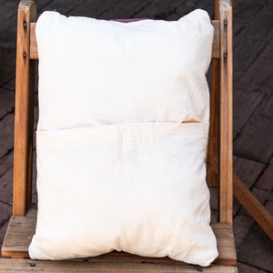 May include: A white pillow with a pocket on the back, sitting on a wooden chair. The chair is made of natural wood and has a rustic look.