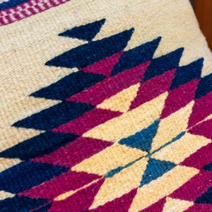 May include: Close-up of a woven pillow with a geometric pattern in shades of blue, pink, and cream.