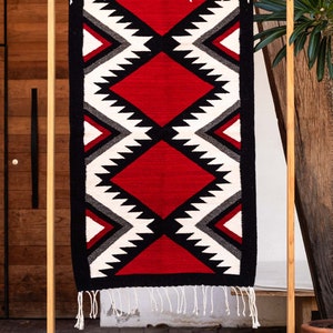 May include: A red, black, white, and gray geometric patterned rug with fringe hanging from the bottom. The rug is displayed on a wooden stand.