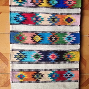 May include: A rectangular, woven rug with a cream-colored background and colorful geometric patterns. The design features horizontal bands of various colors, including blue, green, red, and yellow, with repeating diamond and star-like motifs. The rug has fringed edges.