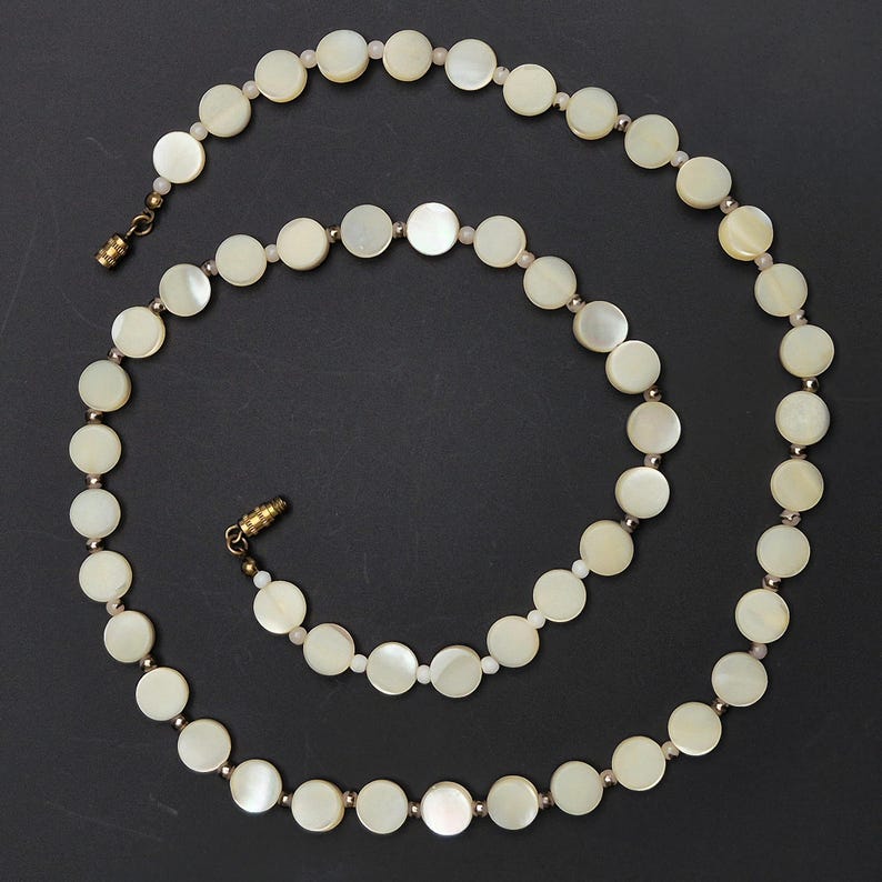May include: A delicate necklace featuring round, off-white mother-of-pearl beads. The beads are connected by small, gold-toned metal links and a matching clasp. The necklace is displayed on a dark background.