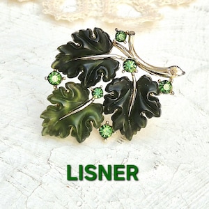 May include: Vintage Lisner brooch featuring a leaf design with dark green leaves and gold-tone accents. The brooch is adorned with green rhinestone accents. The word "LISNER" is printed in green at the bottom of the image.