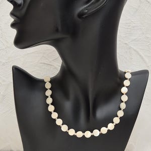 May include: A delicate necklace featuring a strand of round, off-white shell beads. The beads are connected by small, silver-toned links. The necklace is displayed on a black mannequin bust against a white background.