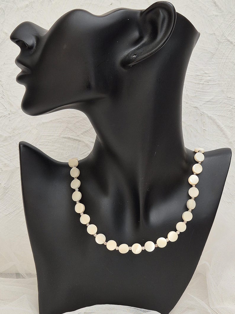 May include: A delicate necklace featuring a strand of round, off-white shell beads. The beads are connected by small, silver-toned links. The necklace is displayed on a black mannequin bust against a white background.