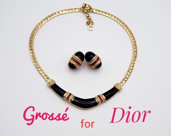 May include: A gold-tone necklace and matching earrings set. The necklace features a black enamel center with red and clear crystal accents. The earrings are black with red and clear crystal accents. The set is from the brand Grosse for Dior.