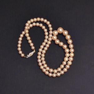 May include: A double strand necklace of peach-colored pearls. The pearls graduate in size, with larger pearls at the center. The necklace is fastened with a silver clasp. The background is a solid black.