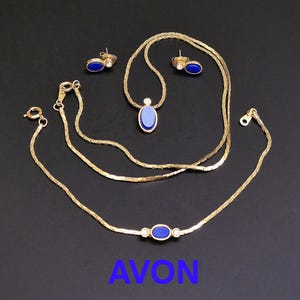 Pode incluir: Um conjunto de joias douradas com colar, pulseira e brincos. Cada peça tem uma pedra oval azul e pequenos detalhes transparentes. A palavra "AVON" é exibida em azul na parte inferior da imagem.