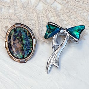 May include: Two vintage brooches. One is an oval-shaped brooch with a green, blue, and brown abalone shell inlay. The other is a silver-toned brooch shaped like a bow with green and blue abalone shell accents.