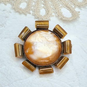 Puede incluir: Broche vintage con una piedra central redonda de color crema, rodeada por un marco de metal bronce. Ocho acentos rectangulares de color ámbar están espaciados alrededor del perímetro, creando un diseño de sol. El broche está sobre un fondo blanco.