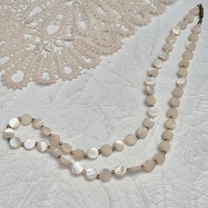 May include: A delicate necklace featuring iridescent, round mother-of-pearl beads. The necklace is strung with small, silver-toned beads between each mother-of-pearl disc. The necklace is displayed on a textured, white surface, with a lace doily in the background.