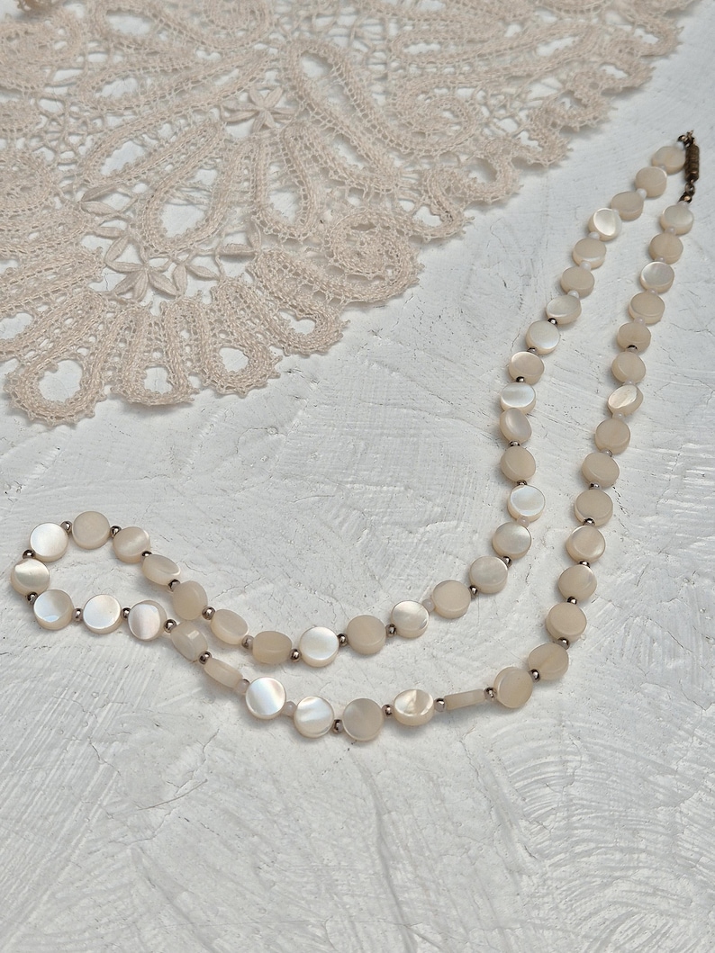May include: A delicate necklace featuring iridescent, round mother-of-pearl beads. The necklace is strung with small, silver-toned beads between each mother-of-pearl disc. The necklace is displayed on a textured, white surface, with a lace doily in the background.