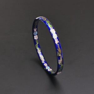 Pode incluir: Uma pulseira circular fina com uma base de esmalte azul royal profundo. A superfície é decorada com um padrão contínuo de delicados motivos florais multicoloridos em tons de rosa, branco e azul claro, acentuados por folhas verdes.