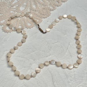 May include: A delicate necklace featuring round, iridescent mother-of-pearl beads. The beads are strung together with small, silver-toned spacers. The necklace is secured with a vintage-style clasp. The jewelry is displayed on a white textured surface.