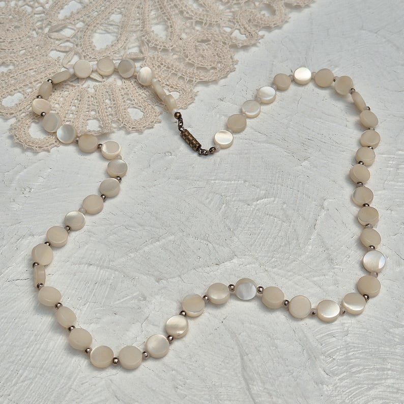 May include: A delicate necklace featuring round, iridescent mother-of-pearl beads. The beads are strung together with small, silver-toned spacers. The necklace is secured with a vintage-style clasp. The jewelry is displayed on a white textured surface.