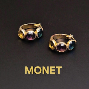 Può includere: Due orecchini a cerchio dorati, ciascuno ornato da tre pietre preziose colorate: giallo, viola e verde acqua. La parola "MONET" è visualizzata in lettere dorate sotto gli orecchini su sfondo nero.
