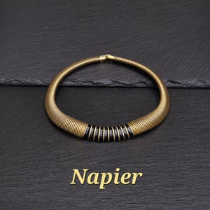 Könnte beinhalten: Eine goldfarbene Metall-Halskette mit geripptem, röhrenförmigem Design und einem Mittelteil aus abwechselnden schwarzen und goldenen Bändern. Die Halskette ist kreisförmig und zeigt den Markennamen Napier in goldenem Text unter dem Schmuckstück.