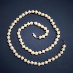 May include: A strand of cream-colored pearls arranged in a spiral on a dark blue background. The necklace features a silver clasp with a small decorative element. The pearls are evenly sized and have a smooth, lustrous appearance.
