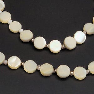 May include: Close-up of two strands of a necklace featuring large, round, off-white mother-of-pearl beads. The beads are interspersed with smaller, iridescent beads and silver-colored spacers. The background is a solid black.