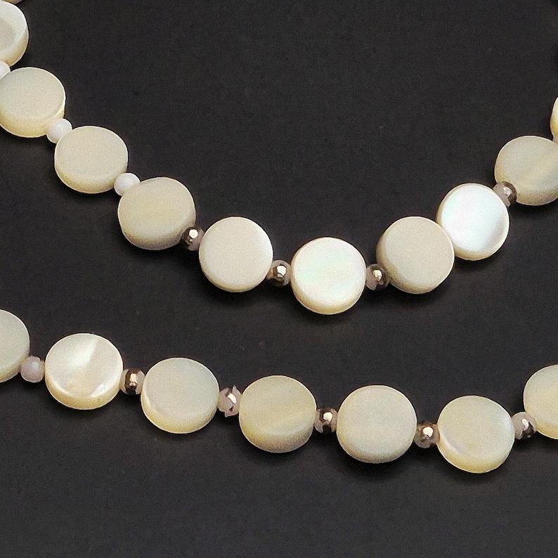 May include: Close-up of two strands of a necklace featuring large, round, off-white mother-of-pearl beads. The beads are interspersed with smaller, iridescent beads and silver-colored spacers. The background is a solid black.