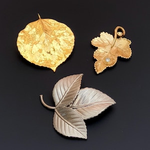 Op de afbeelding: Drie gouden blad broche, één met een enkel blad, één met een cluster van bladeren en één met een enkel blad met een klein diamant accent.