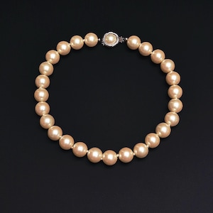 May include: A strand of cream-colored faux pearls, arranged in a circular shape, against a black background. The necklace features a silver-toned clasp with a single pearl at the center. The pearls are evenly spaced and have a smooth, lustrous finish.