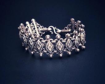Puede incluir: Un brazalete de plata con una cadena delicada y diseños intrincados en forma de diamante. El brazalete tiene un cierre de broche.