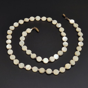 May include: A cream-colored necklace composed of round, flat beads. The beads are strung together with small, gold-toned connectors. The necklace is displayed on a black background, showcasing its elegant design. The clasp is also gold-toned.
