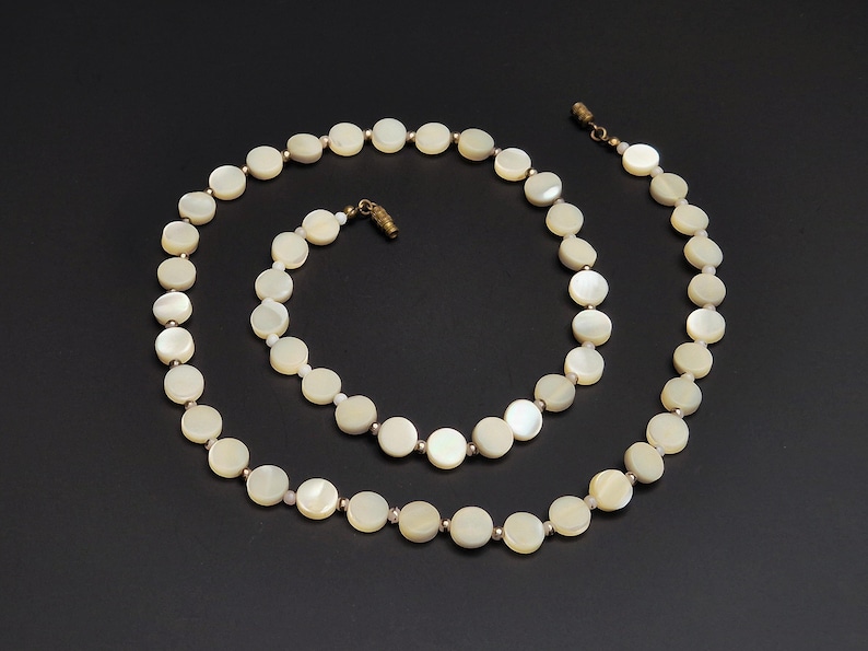 May include: A cream-colored necklace composed of round, flat beads. The beads are strung together with small, gold-toned connectors. The necklace is displayed on a black background, showcasing its elegant design. The clasp is also gold-toned.