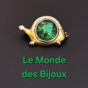 May include: A gold-toned turtle brooch with a green center and a border of clear crystals. The turtle's shell has a circular green design. The brooch has a pin clasp on the back.