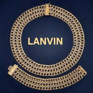Puede incluir: Juego de collar y pulsera dorados con diseño de eslabones de cadena. El collar es circular y la pulsera es una pieza a juego. La palabra "LANVIN" se muestra en letras doradas sobre un fondo azul oscuro.