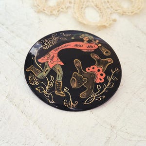 Pode incluir: Broche redonda preta com um design pintado à mão. Apresenta uma figura com túnica vermelha tocando uma trompa, um urso e detalhes florais dourados. A broche tem um acabamento brilhante.